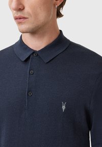 Navyblauwe polo gemaakt van gestructureerde stof, met een klassieke kraag, vier zwarte knopen en een klein grijs geiten-schedel geborduurd op de borst.