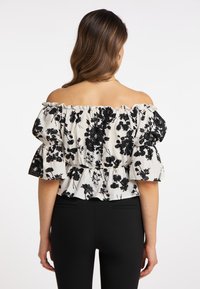 Schulterfreies Oberteil in Weiß mit schwarzem Blumenmuster, Rüschensäumen und taillierter Passform. Kombiniert mit schwarzen Hosen.