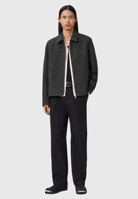 Donkergrijze wolblend jas met een kraag, een rits aan de voorkant en twee zijzakken, gedragen over een wit shirt en zwarte broek. Soepele textuur.