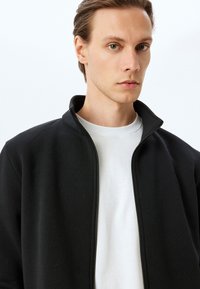 Giacca nera con zip e colletto alto, realizzata in tessuto morbido. Indossata sopra una maglietta bianca, caratterizzata da una texture liscia e un design minimalista.