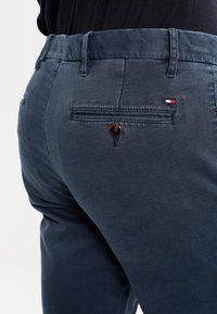 Mörkblå bomullsbukser med en bakficka, dekorativ sömnad och en märkeslogotyp i rött, vitt och blått.