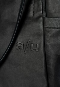 Close-up of a black fabric bag with stitched "a/u" logo and visible seams and texture on the surface.