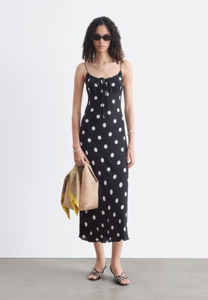 Frau mit lockigem Haar, die schwarze Sonnenbrille und ein schwarzes ärmelloses Kleid mit Punkten trägt, hält eine gewebte Tasche mit einem gelben Schal.