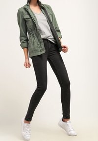 Oliwkowa kurtka robocza, szary t-shirt, czarne dopasowane dżinsy i białe sneakersy. Kurtka z kieszeniami na piersi i podwiniętymi rękawami.