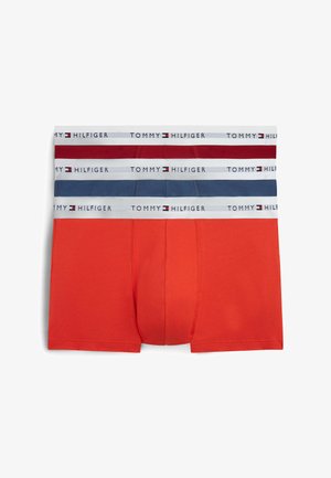 Hombre sin camiseta con los brazos cruzados, lleva bóxers rojos de Tommy Hilfiger sobre un fondo blanco liso.