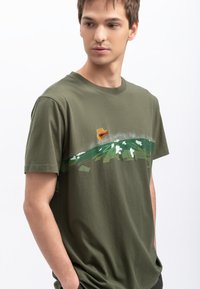 Groene katoenen t-shirt met een grafische print van een landschap met een berg, wolken en een vliegtuig. Korte mouwen en een ronde hals.
