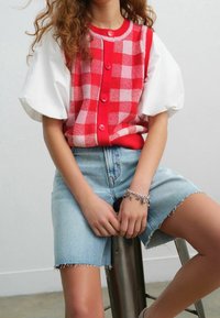 Kobieta siedząca na metalowym stołku, ubrana w czerwoną i różową w kratkę kamizelkę zapinaną na guziki, białą bluzkę z bufiastymi rękawami, jasne dżinsowe szorty oraz bransoletkę z charmsami.