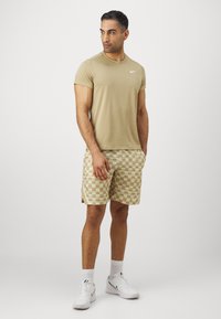 Beige trenings T-skjorte med korte ermer og hvit logo, kombinert med beige rutete shorts. Brukt med hvite joggesko og sokker.