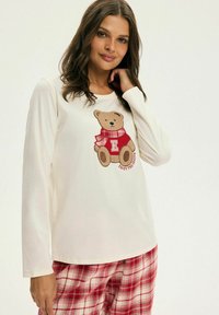 Kremowa koszula z długim rękawem z grafiką misia w czerwonym szaliku. Ma okrągły dekolt. Połączona z czerwono-białymi, kratkowanymi spodniami do piżamy.