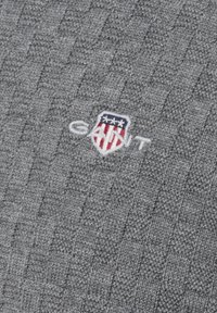 Szary dzianinowy sweter z tkanym wzorem, z małym haftowanym logo w białym, czerwonym i niebieskim kolorze, umieszczonym blisko środka.