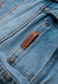Gros plan sur un tissu en denim bleu clair avec des coutures orange, des boutons en métal, et une étiquette en cuir marron sur une poche.