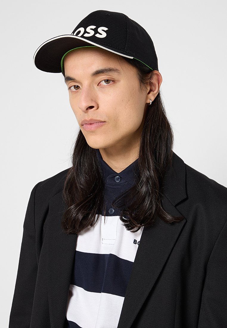 Casquette noire avec le logo "BOSS" en blanc, portée avec un polo rayé bleu marine et blanc et un blazer noir. Cheveux longs et raides visibles.