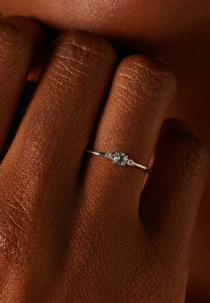 Bague en argent mettant en valeur une pierre précieuse bleu pâle au centre, avec deux petites accents de chaque côté, le tout sur un fond en peau brun texturé.