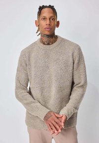 Homme avec tatouages au cou et aux mains, portant un pull beige texturé et un pantalon clair, debout les mains posées doucement devant lui