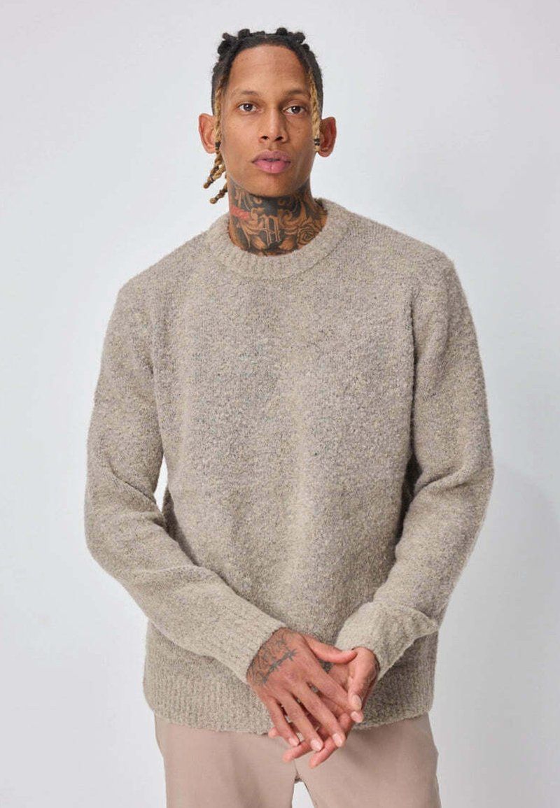 Homme avec tatouages au cou et aux mains, portant un pull beige texturé et un pantalon clair, debout les mains posées doucement devant lui