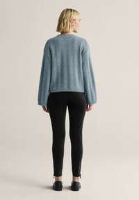 Vaaleansininen neulottu villapaita, jossa on palmikkokuvio, lyhyt malli, leveät hihat, päällä mustien istuvien housujen kanssa, nähtynä takaa.