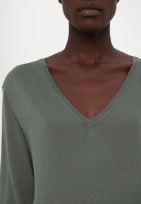 Gros plan d'une personne portant un haut vert désaturé à manches longues et col en V, montrant le cou, les clavicules et le haut de la poitrine.