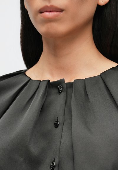 Schwarze, strukturierte Bluse mit rundem Ausschnitt, plissiertem Detail und dekorativen Knopfakzenten entlang der Vorderseite. Glatte Stoffoberfläche.