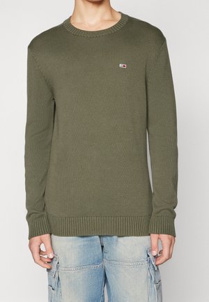 Une jeune personne porte un pull en maille vert olive avec un petit logo sur la poitrine et un jean cargo bleu clair, debout devant un fond uni.