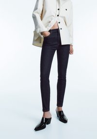 Ciężkie niebieskie dżinsy skinny wykonane z denimu, połączone z kremową, oversize'ową koszulą i czarnymi, lakierowanymi mule na blokowym obcasie.