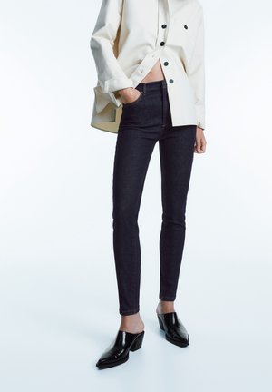 Donkerblauwe skinny jeans van denim, gecombineerd met een crèmekleurig oversized shirt en zwarte gepatineerde leren muiltjes met een blokhak.