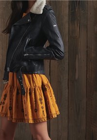 Czarna skórzana kurtka motocyklowa z kołnierzem z baranka, srebrnym zamkiem błyskawicznym i bocznymi kieszeniami; w połączeniu z pomarańczową spódnicą w kształcie dzwonu z haftem.