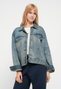 Ανοιχτόγαλαν τζιν μπουφάν με κουμπώματα, δύο τσέπες στο στήθος και μανίκια που έχουν γυριστεί. Διαθέτει ελαφριά ξεθωριάσματα και αντίθετη ραφή.