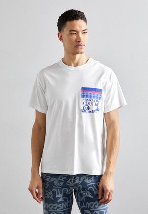 T-shirt bianca in cotone con taschino stampato colorato, dettagliato in blu e rosso, che mostra il testo e il design grafico "VERSACE JEANS COUTURE".