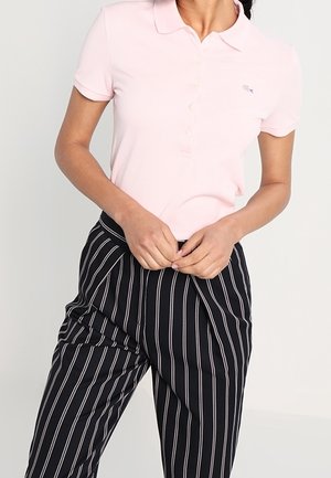 Person wearing a light pink short-sleeve polo shirt and high-waisted black pants with vertical white stripes, hands clasped at waist.