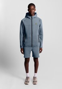 Felpa con cappuccio azzurro chiaro, con zip frontale, tasche laterali e un logo giallo. Abbinata a pantaloni corti blu coordinati e a sneakers neutre.