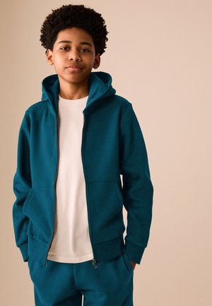 Un adolescente luce una sudadera con capucha de color verde azulado y cremallera, con pantalones a juego sobre una camiseta blanca, delante de un fondo beige liso.