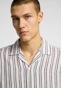 Camisa de manga corta hecha de tela ligera, con rayas verticales en blanco, beige y gris oscuro, con cuello acanalado y botón.