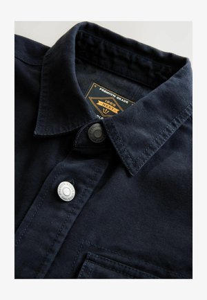 Laivastonsininen puuvillapaita, jossa on napillinen kaulus, hopeiset napit ja rintatasku, teksturoitu kangas ja kudottu etiketti.