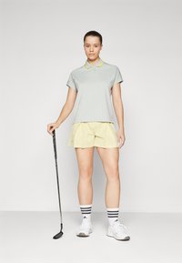 Világosszürke pólóing sárga gallérral, sárga redőzött rövidnadrággal párosítva. Fehér sportcipő és csíkos zokni. Golfütőt tart a kezében.
