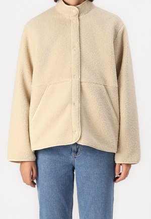 Person trägt eine beige Sherpa-Fleecejacke mit Druckknopfverschluss und Vorderentaschen, kombiniert mit blauen Jeans.