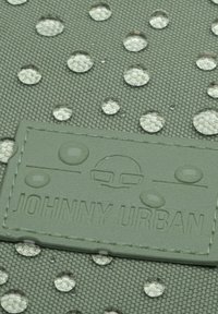 Zöld textúrájú anyag átlátszó vízcseppekkel és a "JOHNNY URBAN" feliratozott logóval. Kör alakú cseppek mintázata látható.