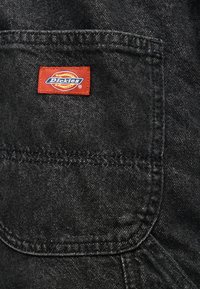 Μαύρη τσέπη από τζιν με μια κόκκινη ορθογώνια ετικέτα της μάρκας Dickies ραμμένη κοντά στο κέντρο του επάνω μέρους της τσέπης.