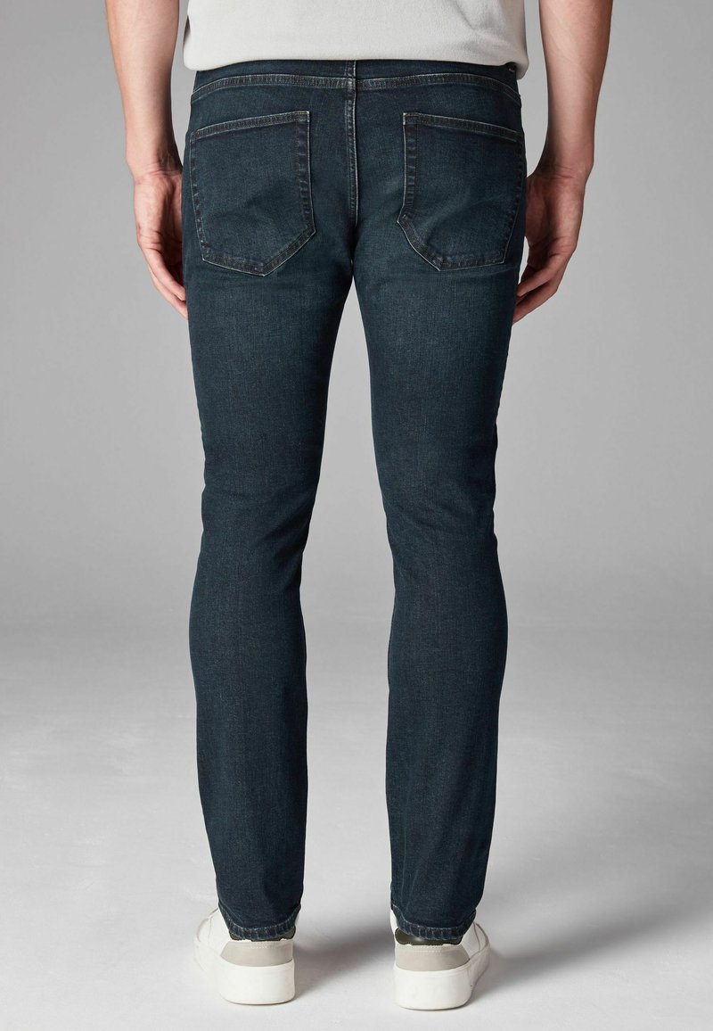Hombre vestido con jeans ajustados de color azul oscuro y zapatillas blancas, mostrado desde atrás sobre un fondo gris liso.