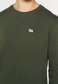 Sudadera verde de algodón con cuello redondo, con un acabado acanalado en el cuello y un pequeño logo blanco de "Lee" en el pecho. La tela parece suave y con textura.