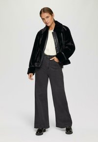 Schwarze Kunstfelljacke mit einem breiten Kragen, kombiniert mit hochgeschnittenen, weit geschnittenen, dunkelgrauen Jeans, die dezente Verzierungen aufweisen. Schwarze Schuhe.