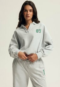 Szary sweatshirt i dopasowane joggery wykonane z miękkiego materiału. Sweatshirt ma kołnierzyk i ozdobiony jest numerem "89" w różowym i zielonym kolorze.