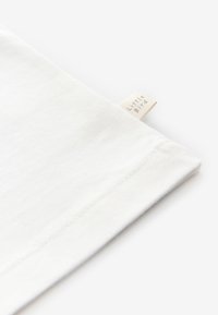 Balta kokvilnas T-krekls ar apakšmala etiķeti "Little Bird." Vienkāršs dizains ar gludu tekstūru un taisnām šuvēm gar malām.