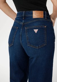 Mörkblå jeans i denim med hög midja, med en läderlabel på baksidan och ett triangelformigt logomärke på fickan.