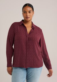 Camisa burdeos de botones con un patrón texturizado, mangas largas y cuello clásico. Combinada con jeans de mezclilla de color azul claro.