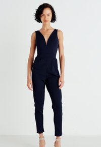 Mørkeblå jumpsuit med dyp V-hals, tettsittende design og smale ben. Laget av glatt stoff uten synlige mønstre eller dekorasjoner.
