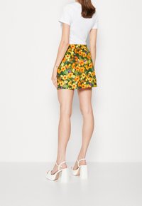 Falda corta floral con fondo verde, que presenta vibrantes flores amarillas, naranjas y moradas. Combinada con sandalias de tacón bloque blancas.