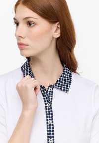 Polo bianco con colletto navy a fantasia con forme tonde, chiusura frontale con bottoni e texture liscia. Design semplice e moderno.