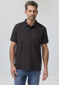 Homme aux cheveux courts et foncés avec une barbe, portant un polo noir et un jean bleu, debout devant un fond gris clair uni.