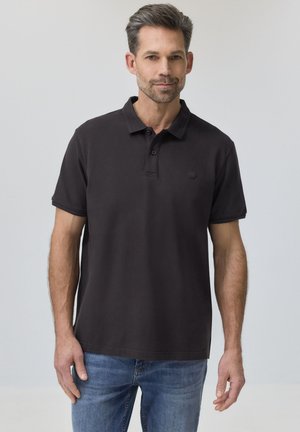 Homme aux cheveux courts et foncés avec une barbe, portant un polo noir et un jean bleu, debout devant un fond gris clair uni.