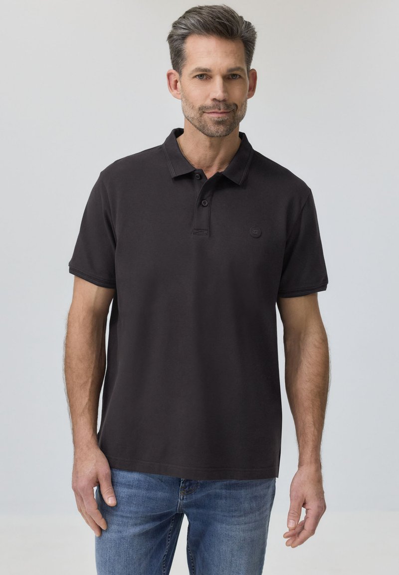 Homme aux cheveux courts et foncés avec une barbe, portant un polo noir et un jean bleu, debout devant un fond gris clair uni.